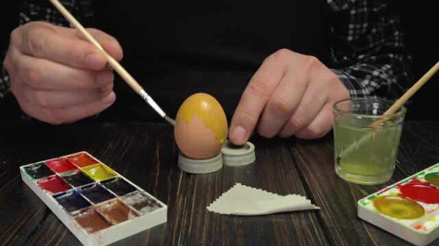 Person painting decorative patterns on an egg using a fine brush, with watercolor palette and glass of water visible on a wooden table