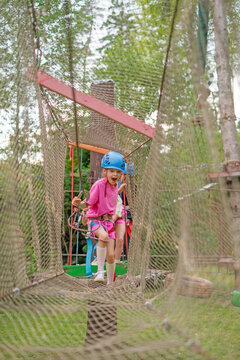 A mischievous girl in a sports helmet and safety equipment is climbing a rope course through a stretched net.