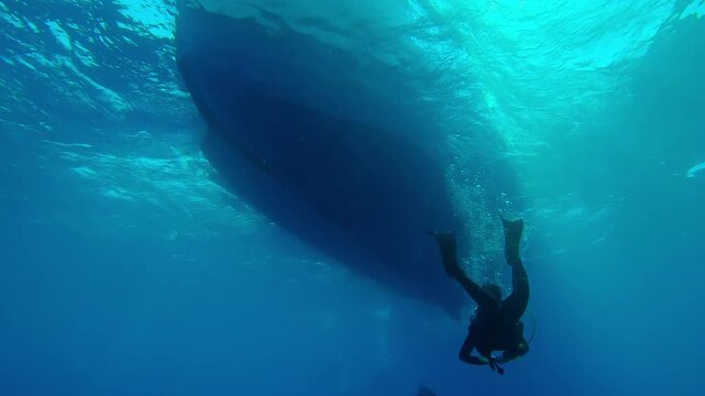 Scuba divers swim in turquoise water towards ramp passing under bottom of diving boat on sunny day in back light, Slow motion of divers returning underwater to diving safari boat in backlit