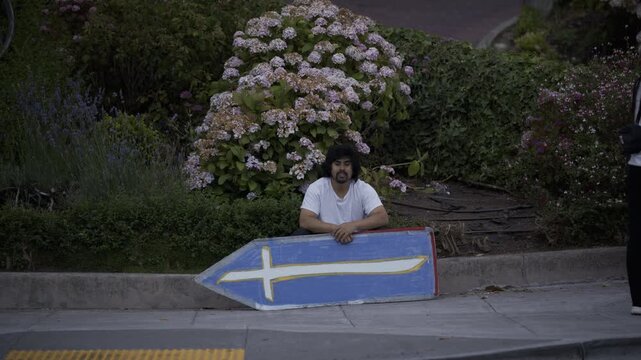 Man Leaning On Blue Cross Board At Sidewalk Near Plants In San Francisco