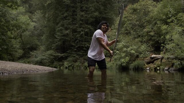 Man Swinging Painted Board Across Surface Of Shallow River In Forest 