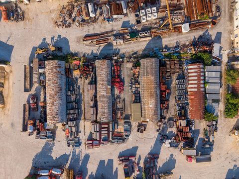 Aerial view of industrial storage yard with arched-roof warehouses, stacks of metal pipes, construction materials, and heavy equipment on a gravel lot in Ravadinovo, Burgas, Bulgaria.