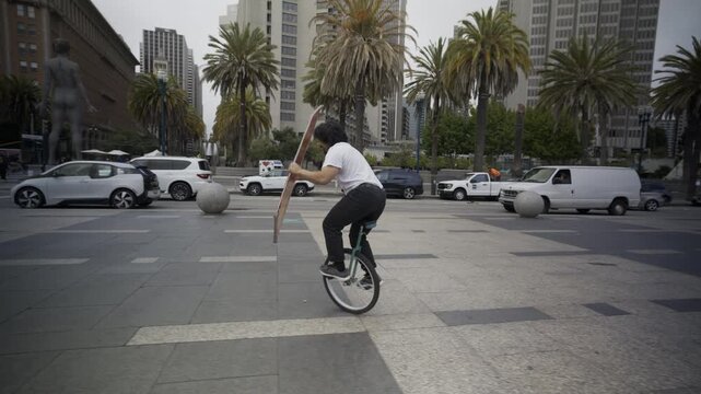 Man Riding Unicycle While Carrying Large Board In Downtown Plaza Of San Francisco