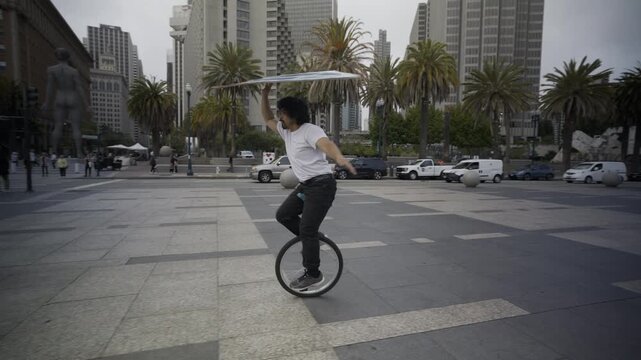 Man Riding Unicycle While Spinning Painted Board In San Francisco Plaza - Steady Cam Shot