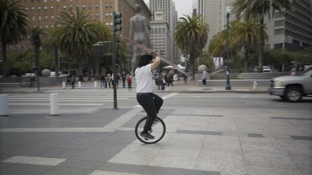 Unicyclist Balancing And Spinning Long Board Overhead While Riding Around Downtown San Francisco Plaza