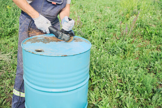 Worker wearing gloves grinding blue metal barrel with an angle grinder, creating sparks outdoors