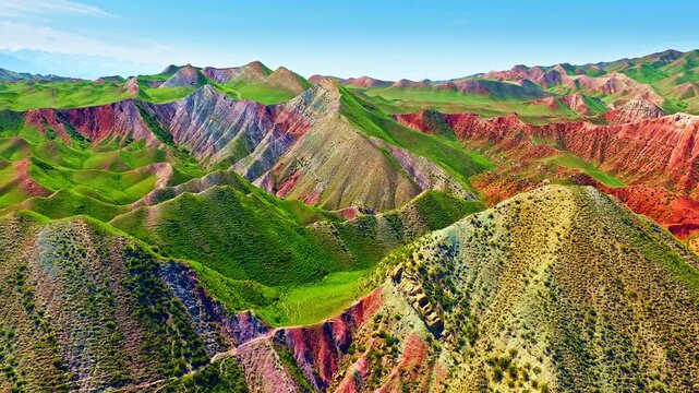 Spectacular aerial view of colorful Danxia landform mountains in Xinjiang, China, showcasing vibrant red, green, and yellow rock layers under a clear blue sky.