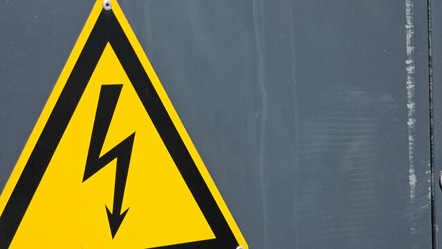 Yellow triangular high voltage warning sign with black lightning bolt symbol attached to a weathered grey metal panel. Industrial electrical safety concept and hazard protection

