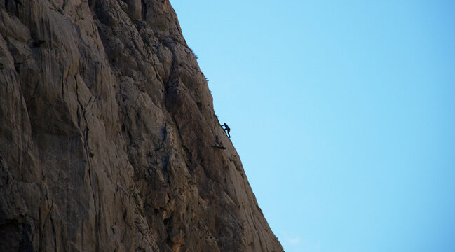 A lone rock climber ascends a steep mountain cliff under a clear blue sky. Adventure, determination, courage, and endurance captured in a dramatic outdoor landscape. Perfect for concepts of challenge.