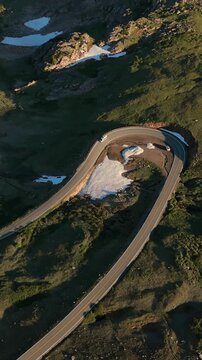 Vertical Screen: Beartooth Highway curves through Montana USA mountain landscape with melting snow patches and alpine tundra. Cars navigate the scenic switchback road at golden hour.