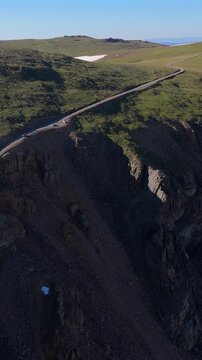 Vertical Screen: Aerial drone footage captures winding Beartooth Highway cutting through rugged alpine landscape with dramatic rocky cliffs, lush green tundra, snow patches.