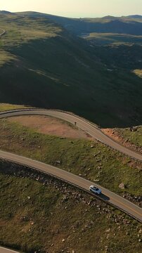Vertical Screen: White SUV navigates serpentine curves of Beartooth Highway in Montana, USA. Aerial drone view captures alpine tundra landscape with dramatic switchbacks at golden hour.