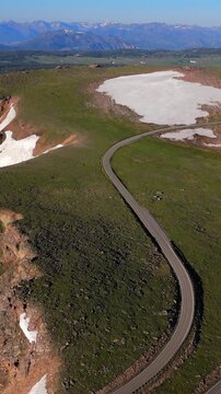 Vertical Screen: Beartooth Highway curves through green alpine meadows with melting snow patches in Montana USA. Distant mountain ranges under clear blue sky create stunning scenic vista.