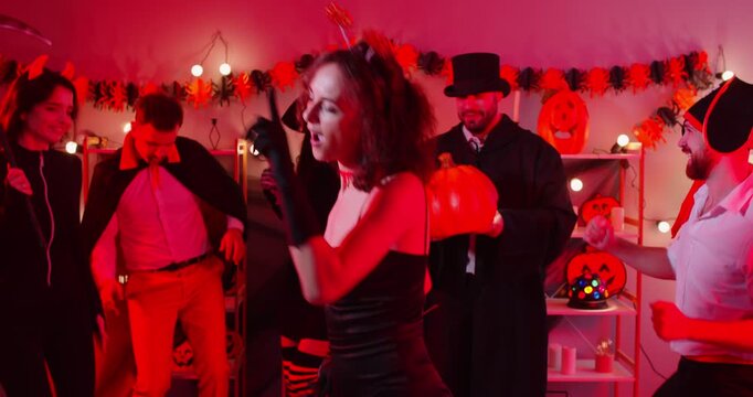 Happy Halloween holiday demon devil woman in black dress dancing with friends wearing costumes for dress ups party night event, playing spooky horror cosplay look, garland red light decor atmosphere