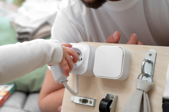 Close-up of a two-year-old toddler inserting a plug into a practice socket on a wooden Montessori busy board while the father watches. Concept of safe learning, early development and fine motor skills