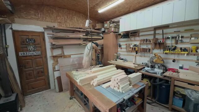 Anonymous handyman in coveralls using heavy electric drill with hole saw bit to cut circular shapes in plywood panel on workbench inside cluttered workshop during handheld wide angle sweeping arc shot