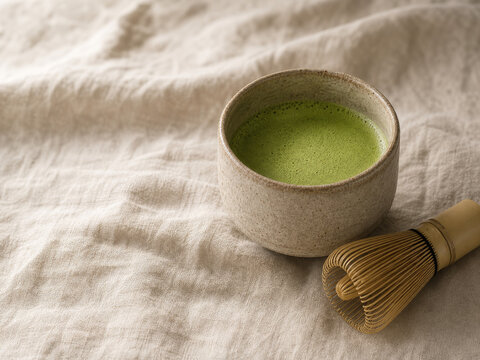 Close up of a textured raw clay chawan bowl filled with vibrant green matcha tea on a wrinkled natural linen fabric background with traditional bamboo chasen whisk.