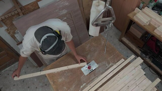 High angle top down overhead view of anonymous carpenter in protective gear shaping long wooden plank on router table milling machine in dusty workshop during authentic documentary handheld shot