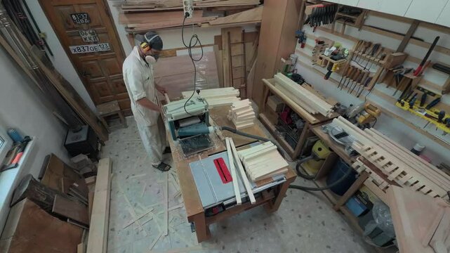 High Angle Wide Shot Of Unrecognizable Solo Carpenter In Earmuffs Operating Thickness Planer Machine Inside Highly Cluttered Woodworking Workshop With Scattered Timber Scraps And Wall Tool Racks
