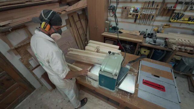 Over The Shoulder Rear View Of Unrecognizable Carpenter In Protective Earmuffs Processing Timber Boards With Thickness Planer On Workbench Inside Messy Local Woodworking Studio Wide Angle Shot