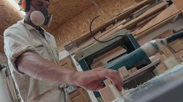 Professional woodworker wearing protective respirator and industrial earmuffs feeding wooden blanks into a thickness planer machine in an authentic garage carpentry workshop during daylight low angle