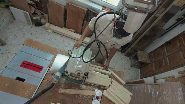Unrecognizable Carpenter In Coveralls And Safety Gear Feeding Timber Planks Into Thickness Planer On Workbench During Busy Workday In Woodworking Studio Handheld High Angle Overhead Documentary