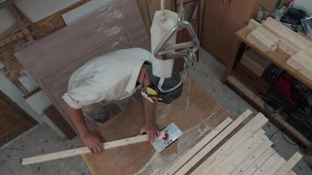 Unrecognizable industrial fabricator in safety coveralls trimming long wooden batten on power router table inside dusty joinery workroom during overhead bird's eye view handheld authentic shot