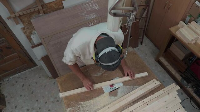 Unrecognizable woodcraft artisan wearing safety apparel shaping thin lumber molding on routing machine inside dusty DIY woodshop during top down overhead authentic documentary handheld shot