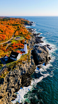 An aerial photograph of a coastal landscape featuring a lighthouse a house and a rocky shoreline with trees displaying autumn colors