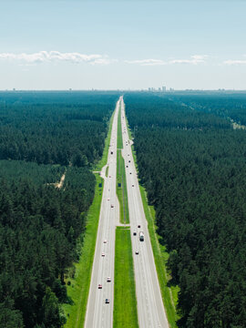 Aerial view of the A2 highway stretching through a vast, dense pine forest with vehicles traveling along the road under a clear blue sky in Riga, Adazi Municipality, Latvia.