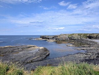 Fototapeta premium Rugged coastline of Ireland featuring dark stratified rocks, a calm blue ocean, and a bright sky.