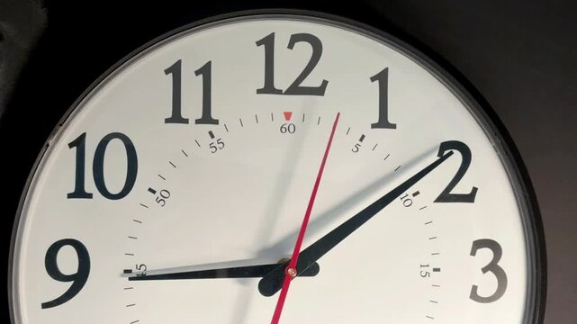 Close-up of round wall clock with white face, bold black numerals, black hour and minute hands, red second hand, slight angle, dark background, soft light, mild shadow, clean minimalist look
