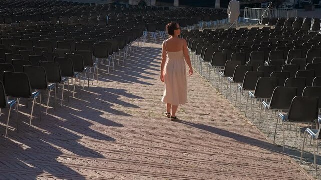 Woman walking through empty chairs in open air venue. Woman walks away from camera between rows of empty black chairs in outdoor venue paved with herringbone bricks, casting long shadows