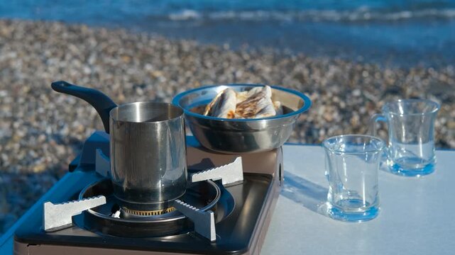 Coffee brewing on portable stove by the sea. Stainless steel coffee pot brewing on a portable gas stove on a beach table, with empty glasses and a plate of cookies nearby, the sea in the background