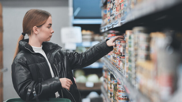 Female shopper selecting canned goods, browsing grocery aisle, comparing product options during weekly shopping trip