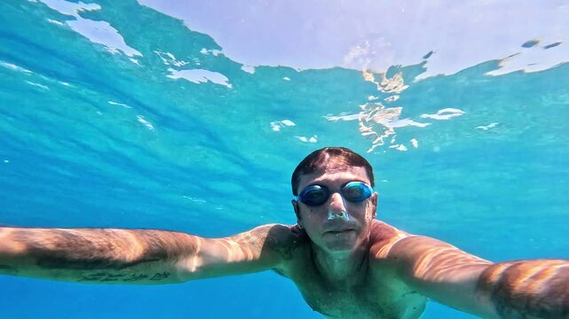 A man swims underwater in the Mediterranean Sea. The man is of Caucasian origin, is forty-two years old, has short hair and has swimming goggles on his face