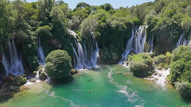 Aerial drone view approaching majestic waterfall with emerald river surrounded by lush green forest in summer