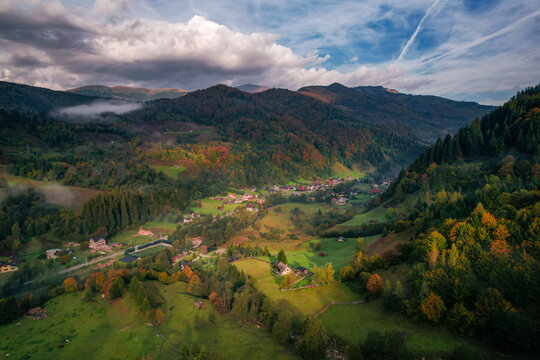 Aerial view of Saticu de Sus nestled in the lush green valley of the Carpathian Mountains with autumn foliage, rolling hills, and scattered houses in Satic, Judetul Arges, Romania.