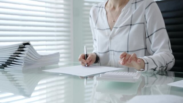 Woman's hands using a calculator and holding a pen, working with financial documents and a pile of papers on a clear desk in an office setup