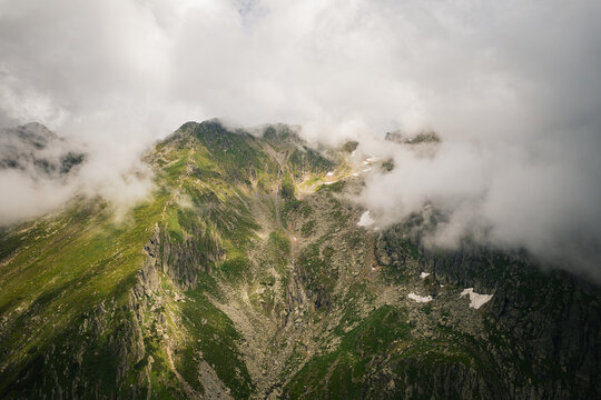 Aerial view of Negoiu Peak in the Fagaras Mountains, featuring rugged rocky slopes, patches of snow, and green vegetation partially obscured by clouds. Porumbacu de Jos, Sibiu, Romania.