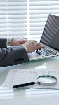 Businessman's hands are actively typing on a laptop keyboard, researching data and analyzing financial documents laid out on a clear desk with a magnifying glass in an office setting