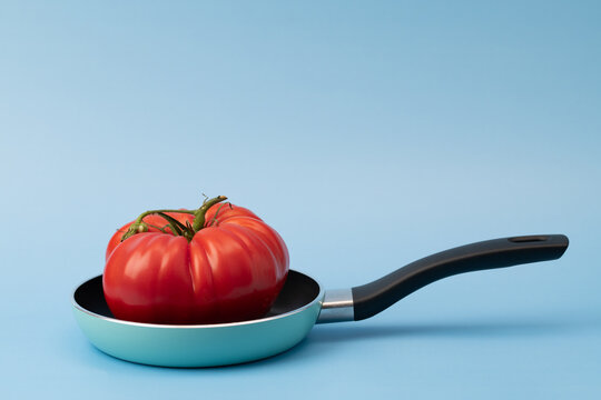 There is a red tomato in a pan. The pan and the tomato are set against a blue background. There is space for text.