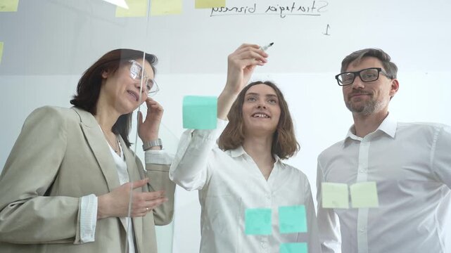 Business professionals collaborating at office meeting, developing strategy and brainstorming ideas on a transparent glass board, working together for project success and innovation