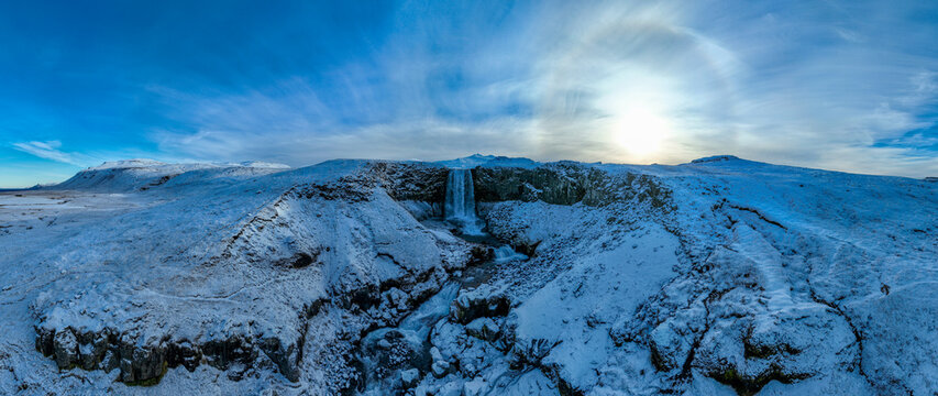 Aerial view of a waterfall cascading down a snow-covered rocky cliff in a vast winter landscape under a bright sun with a halo in the sky Grindavik, Sveitarfelagid Vogar, Iceland.