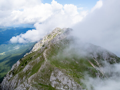 Aerial view of Piatra Craiului Mountains with rocky limestone ridges, green alpine meadows, and drifting clouds in Zarnesti, Brasov, Romania.