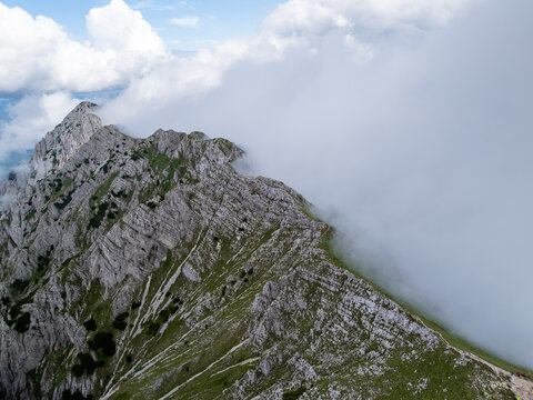 Aerial view of the rugged rocky ridge of the Piatra Craiului Mountains partially obscured by thick clouds and mist above green slopes in Zarnesti, Brasov, Romania.