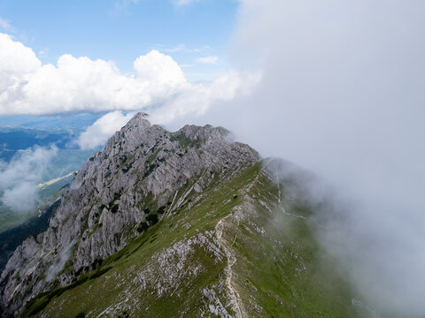 Aerial view of Piatra Craiului Mountains with jagged limestone ridges, green alpine meadows, and clouds rolling over the peaks in Zarnesti, Brasov, Romania.