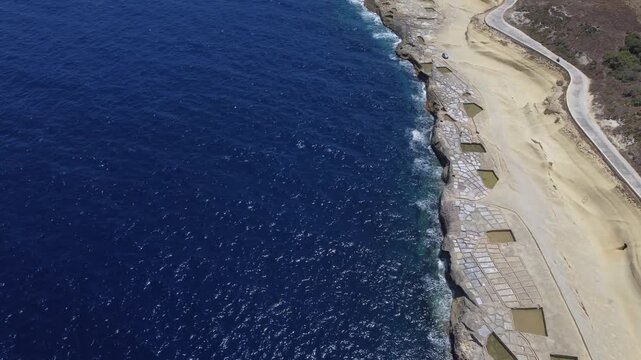 Xwejni s centuries-old salt pans unfold along the rugged coast of Zebbug, Gozo, geometric pools and vivid Mediterranean blues in a sweeping summer Aerial view. High quality 4k footage