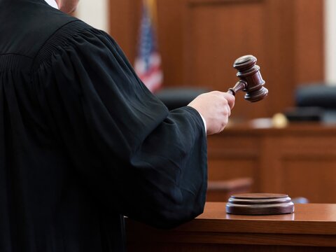 A judge in a black robe prepares to strike a wooden gavel on a sound block in a solemn courtroom.