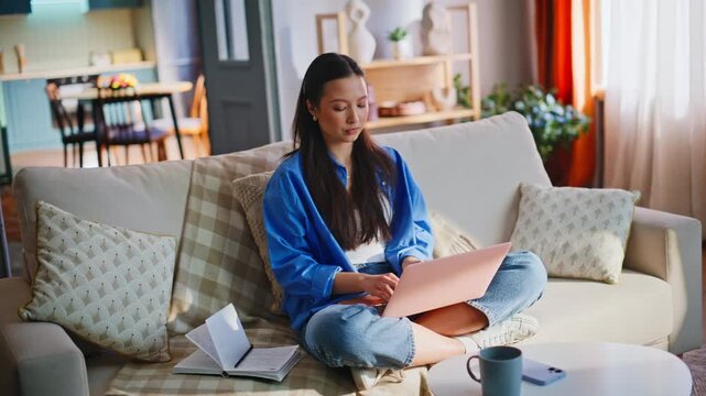 Pensive Asian freelancer pausing work pink laptop sofa. Woman planning ideas home.